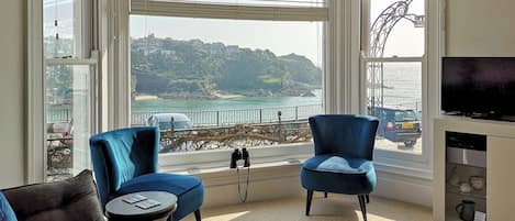 A room with a large bay window, two blue armchairs, a small round table, a flat-screen TV, and a view of a coastal landscape.