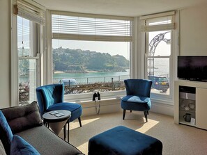 Une pièce dotée d’une grande baie vitrée, de deux fauteuils bleus, d’une petite table ronde, d’un téléviseur à écran plat et offrant une vue sur un paysage côtier.