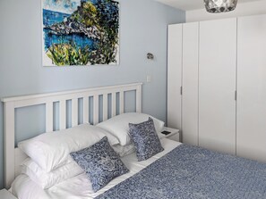 A bedroom with a white bed, patterned pillows, a blue and white bedspread, a white headboard, a white wardrobe, and a painting of a landscape.