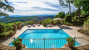 A rectangular pool with a stone deck, surrounded by a metal fence, and overlooking a lush landscape.