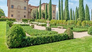 A historic building with a symmetrical garden, tall cypress trees, and a clear sky.
