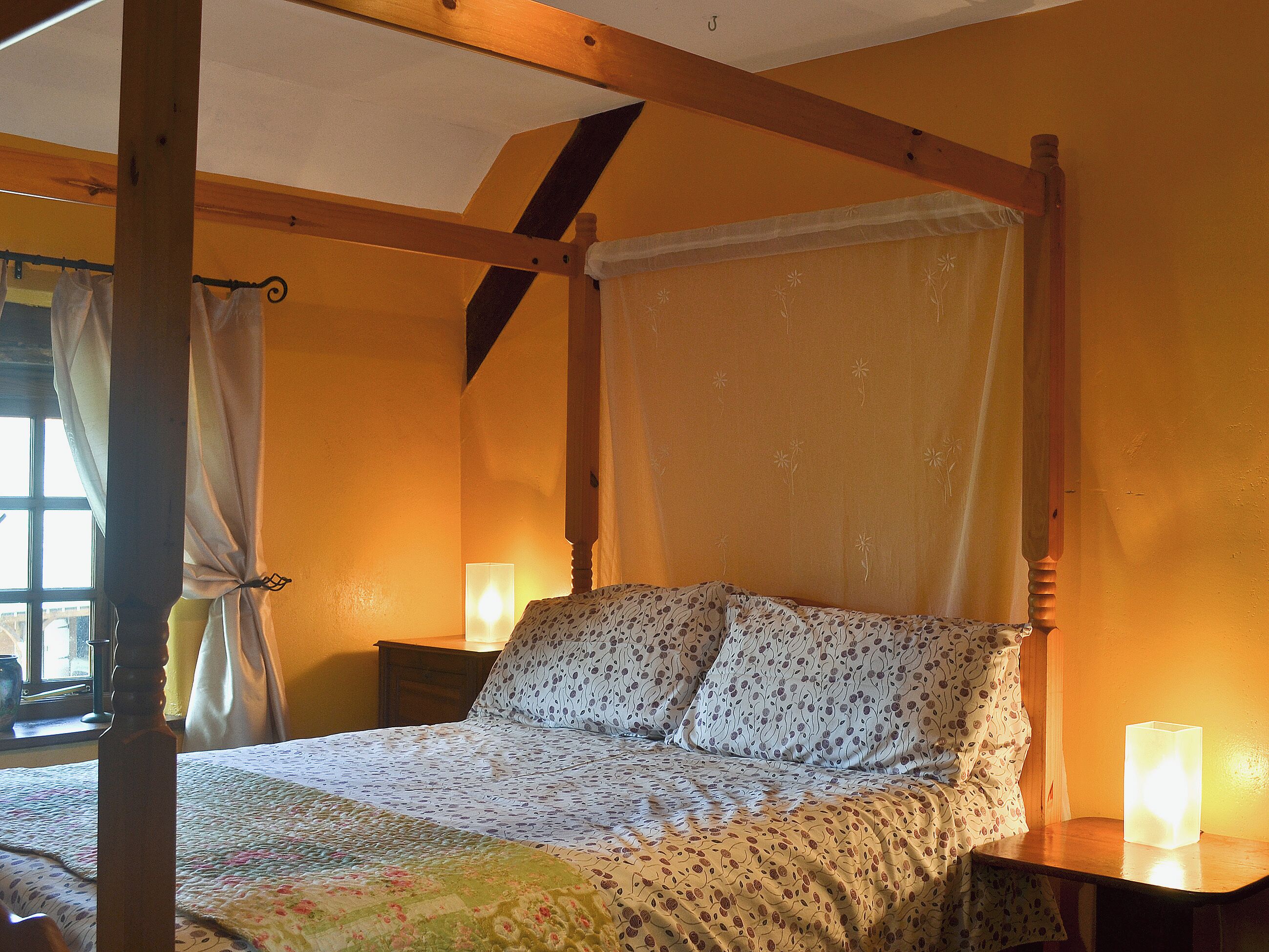 Ein Himmelbett mit Baldachin, ein Nachttisch mit Lampe und ein Fenster mit Vorhängen.