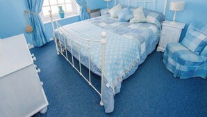 Een slaapkamer met een wit metalen bedframe, een blauw bed met stippen, bijpassende blauwe gordijnen en een blauw vloerkleed.