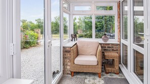 A cozy room with a wicker chair, a small table, and a view of the garden through large windows.