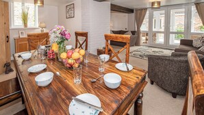 A dining area with a wooden table set for four, surrounded by a living room with a sofa and a view of the outside.