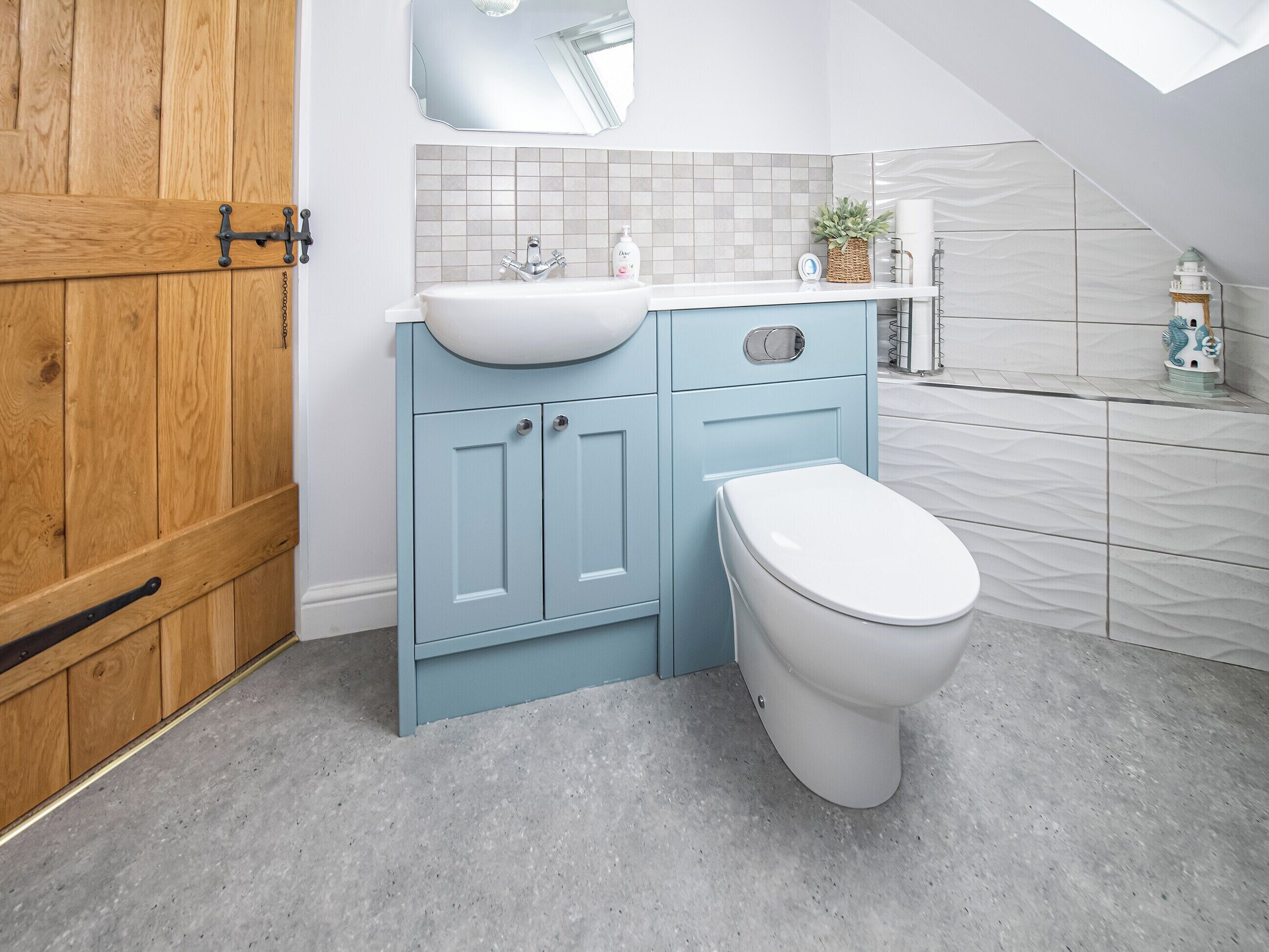 A modern bathroom with a wooden door, a light blue vanity, a white sink, and a toilet.