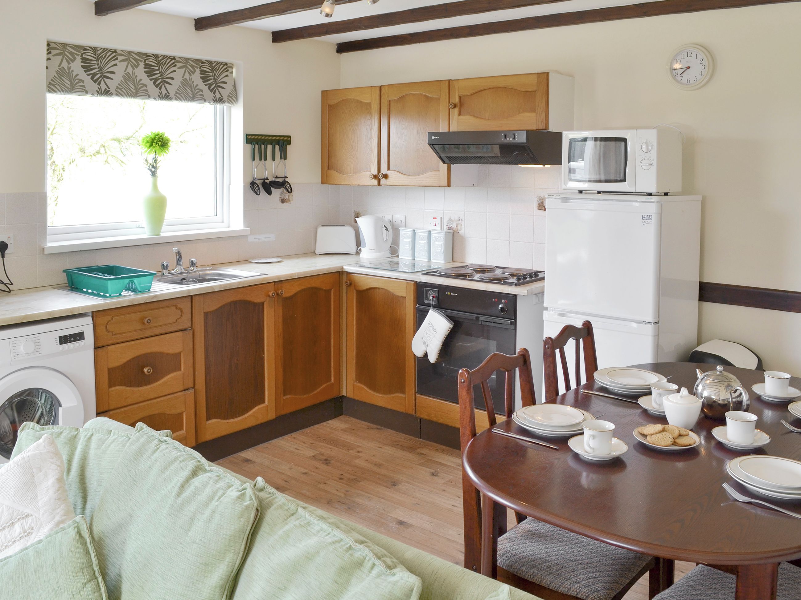A compact kitchen with wooden cabinets, a built-in oven, microwave, and a refrigerator. There's a dining table set with plates, cups, and a kettle.