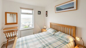 A bedroom with a bed, a chair, a nightstand, a lamp, a window with a view, and a framed picture on the wall.