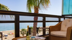 Uma varanda com uma cadeira de vime, um tabuleiro com uma garrafa de vinho, um copo e uma pequena tigela. A vista inclui uma praia e palmeiras.