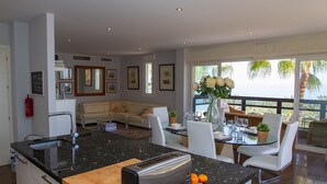 Cozinha moderna com torradeira, pia e área de jantar com mesa e cadeiras. Há uma varanda com vista para palmeiras e o mar.