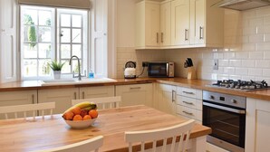 A kitchen with white cabinets, a microwave, an oven, and a wooden table with a bowl of fruit. - No. 1 The Old School (Puddletown, near Dorchester)