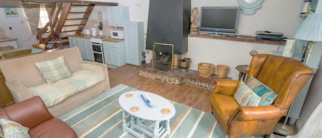 A cozy living room with a sofa, armchair, and a small coffee table. There is a fireplace, a television, and a kitchen area in the background.