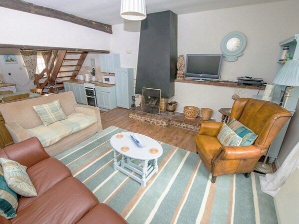 A cozy living room with a sofa, armchair, and a small coffee table. There is a fireplace, a television, and a kitchen area in the background. - Buttercup Cottage - UKC2847 (Blakeney, near Holt)