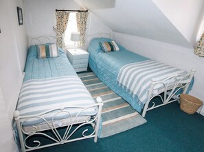 A bedroom with two single beds, a nightstand, a lamp, and a window with curtains. - Buttercup Cottage - UKC2847 (Blakeney, near Holt)