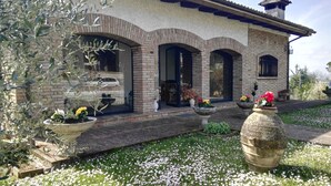 A house with a brick facade, arched doorways, and a tiled roof. There are flower pots on the porch and a lawn with white flowers. - Villa Mery, Holiday house in the hills of the Rimini Riviera, with private pool (Mondaino)
