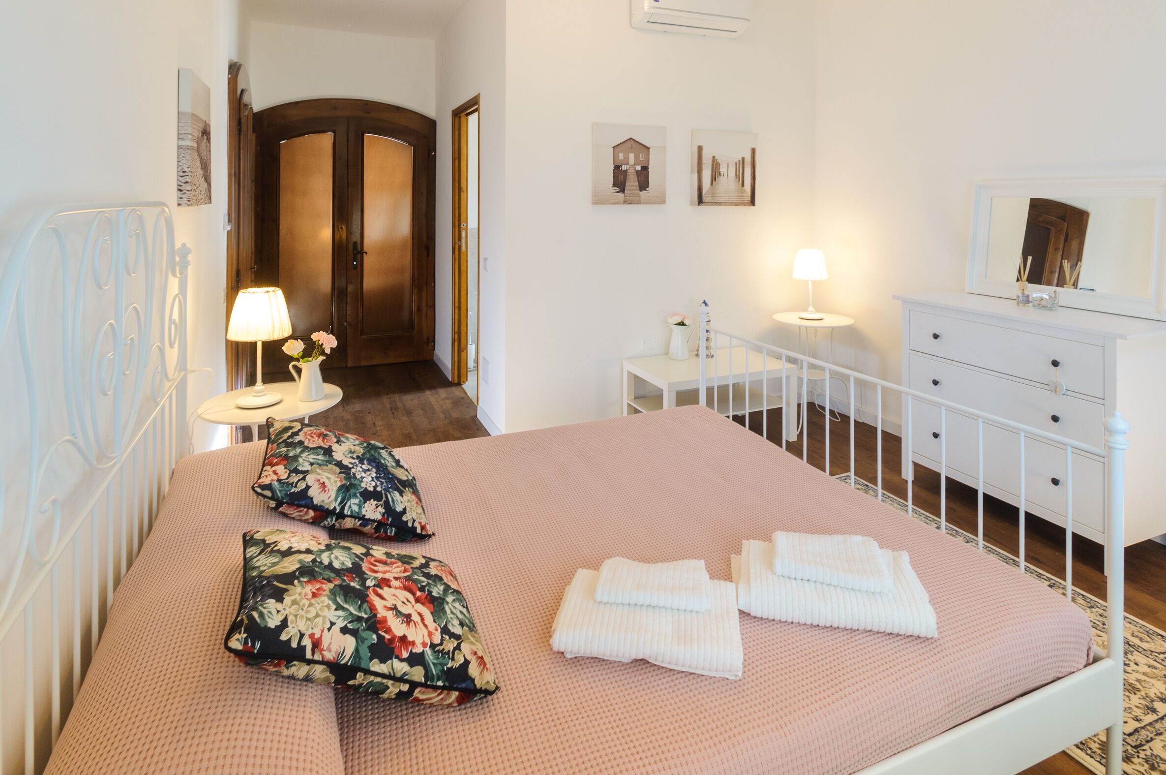 Une chambre à coucher bien rangée, avec un lit, des oreillers, une table de chevet avec une lampe, une commode et une porte donnant sur une autre pièce.