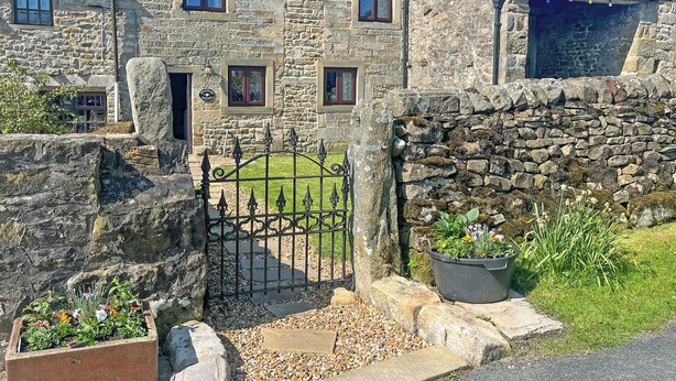 Une charmante maison en pierre avec une entrée fermée par un portail, un petit jardin agrémenté de plantes en pot et un ciel d’un bleu éclatant.