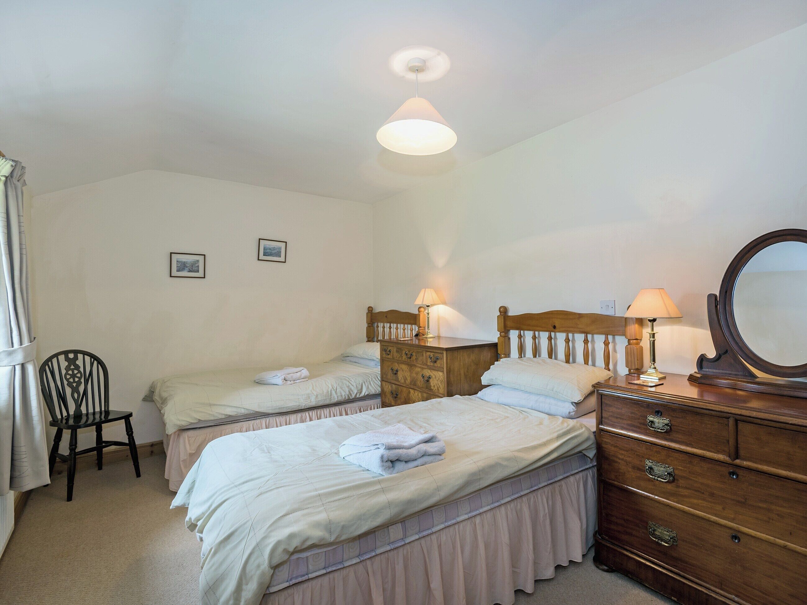 Een slaapkamer met twee enkele bedden, een houten dressoir met lades, een stoel en twee bedlampjes.