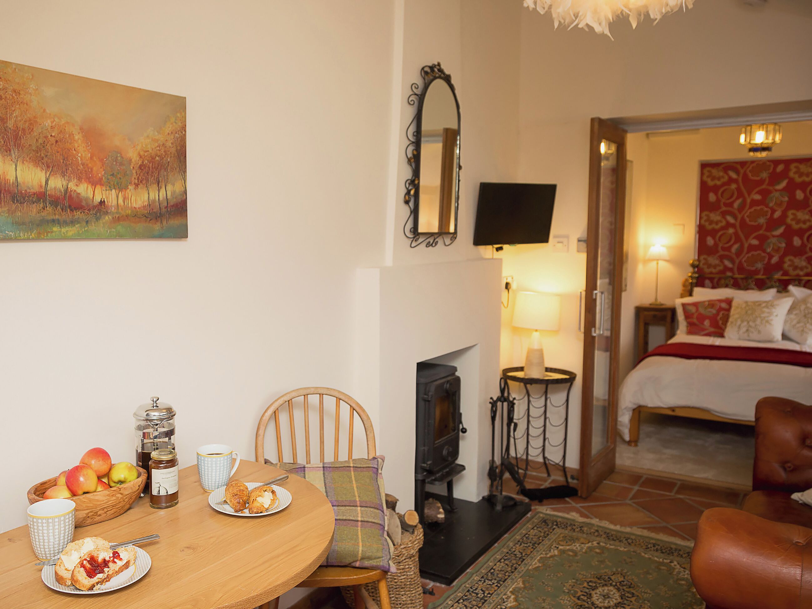 Une pièce chaleureuse avec une table à manger dressée pour le petit-déjeuner, un lit recouvert d’une courtepointe à motifs floraux rouges et une cheminée.
