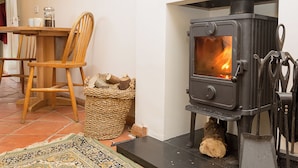 A cozy room with a wood-burning stove, a woven basket of firewood, a wooden chair, and a tiled floor. - Gamekeeper's Cottage (Staple, near Wingham)