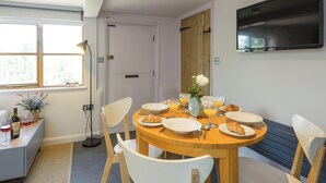 A dining area with a round wooden table, white chairs, and a TV mounted on the wall. - 2 bedroom accommodation in Blythburgh, near Southwold (Blythburgh, near Southwold)