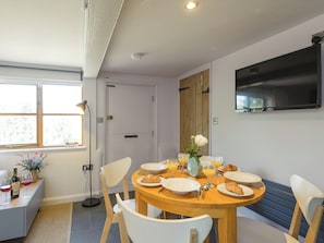 A dining area with a round wooden table, white chairs, and a TV mounted on the wall. - 2 bedroom accommodation in Blythburgh, near Southwold (Blythburgh, near Southwold)
