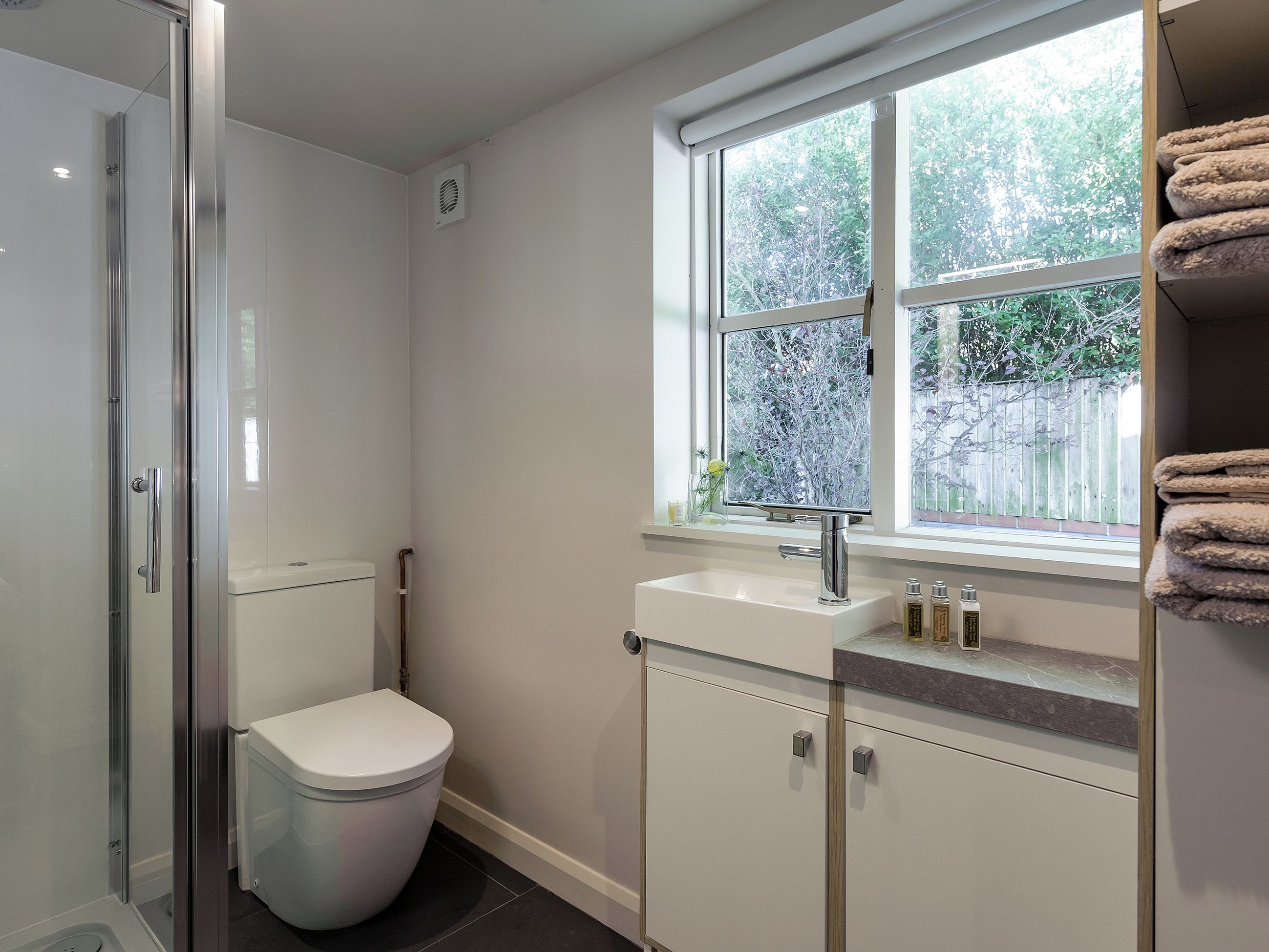 A modern bathroom with a glass shower enclosure, a toilet, a sink with a window above, and a towel rack.