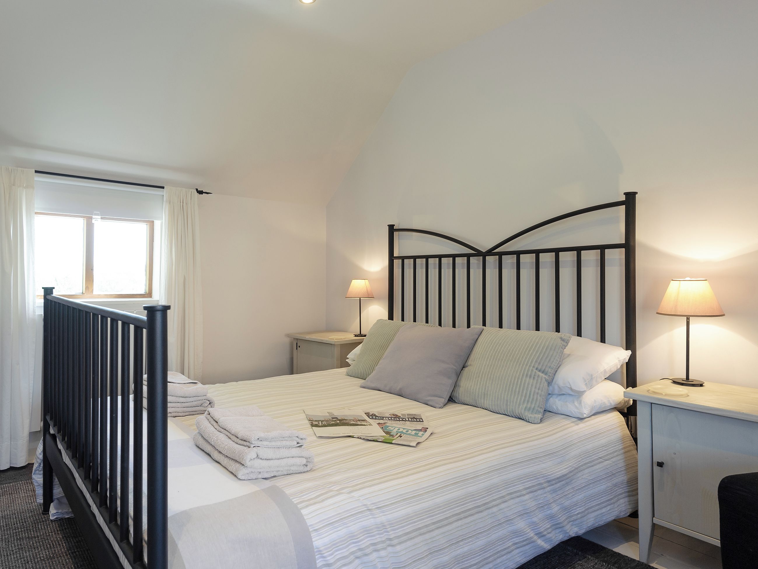 Un dormitorio con una cama, dos mesitas de noche, una mesilla auxiliar, una ventana con cortinas y un radiador.