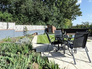 An outdoor patio with a table and chairs, surrounded by a garden with various plants and a brick wall in the background. - 2 bedroom accommodation in Blythburgh, near Southwold (Blythburgh, near Southwold)