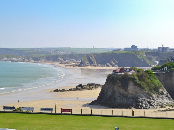 A coastal beach with a grassy area in the foreground, a sandy beach, and a cliff with a building on top.