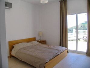 Quarto com cama de madeira, mesa de cabeceira, abajur e vista para uma varanda com guarda-corrimão.