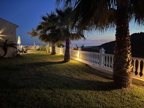 A well-lit outdoor area with a white fence, palm trees, and a building with an umbrella. - Casa Bonita | Authentic Andalusian House (Algarrobo)
