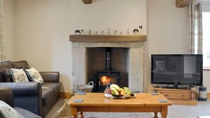 A cozy living room with a stone fireplace, a leather sofa, a wooden coffee table with fruit, and a television. - Little Grans Cottage (Ickornshaw, near Cowling)