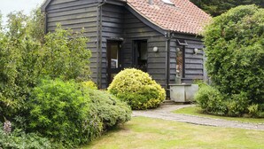 A dark wooden house with a sloped roof, surrounded by lush greenery and a stone pathway leading to the entrance. - 3 bedroom accommodation in Sternfield, near Aldeburgh (Sternfield, near Aldeburgh)