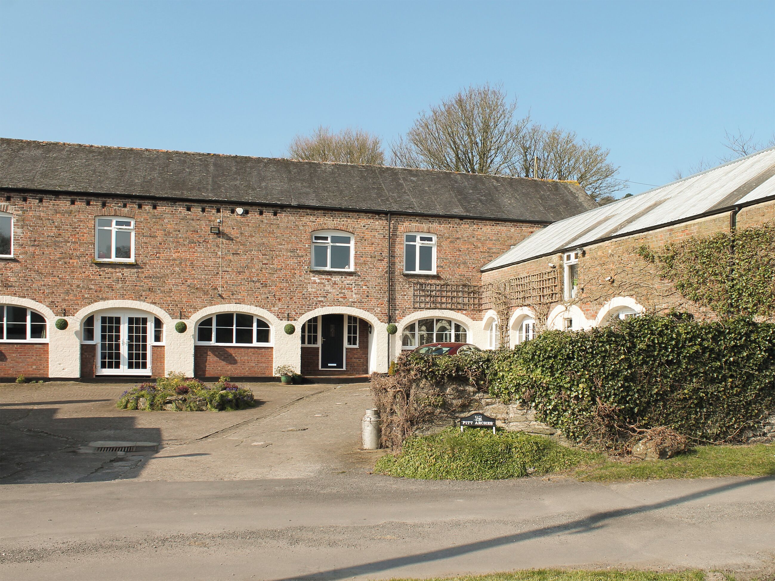 A brick building with arched windows and a door, surrounded by greenery and a clear sky.