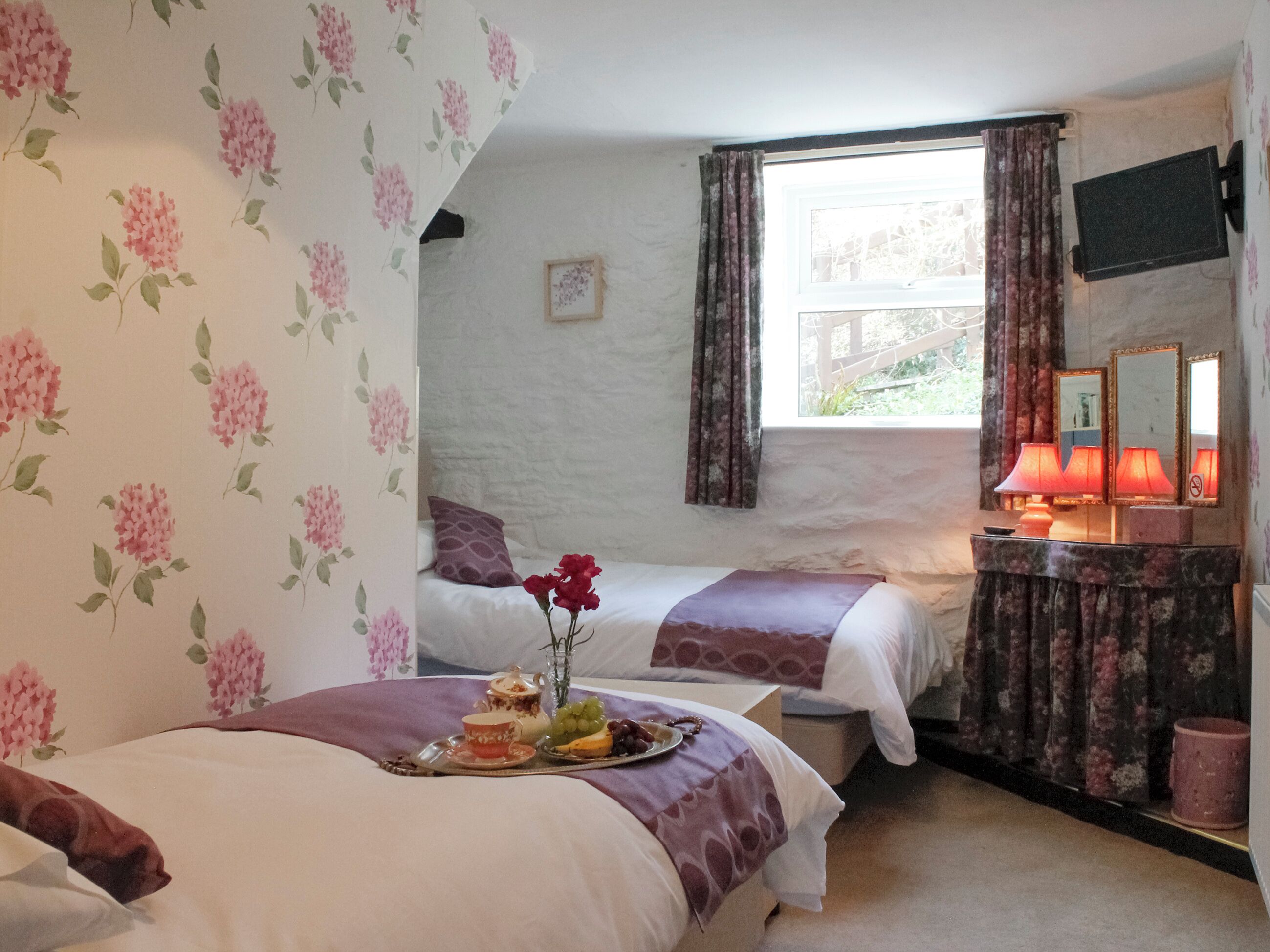 A hotel room with two beds, floral wallpaper, a window with curtains, a TV, a bedside table with a lamp, and a tray with food and drinks.