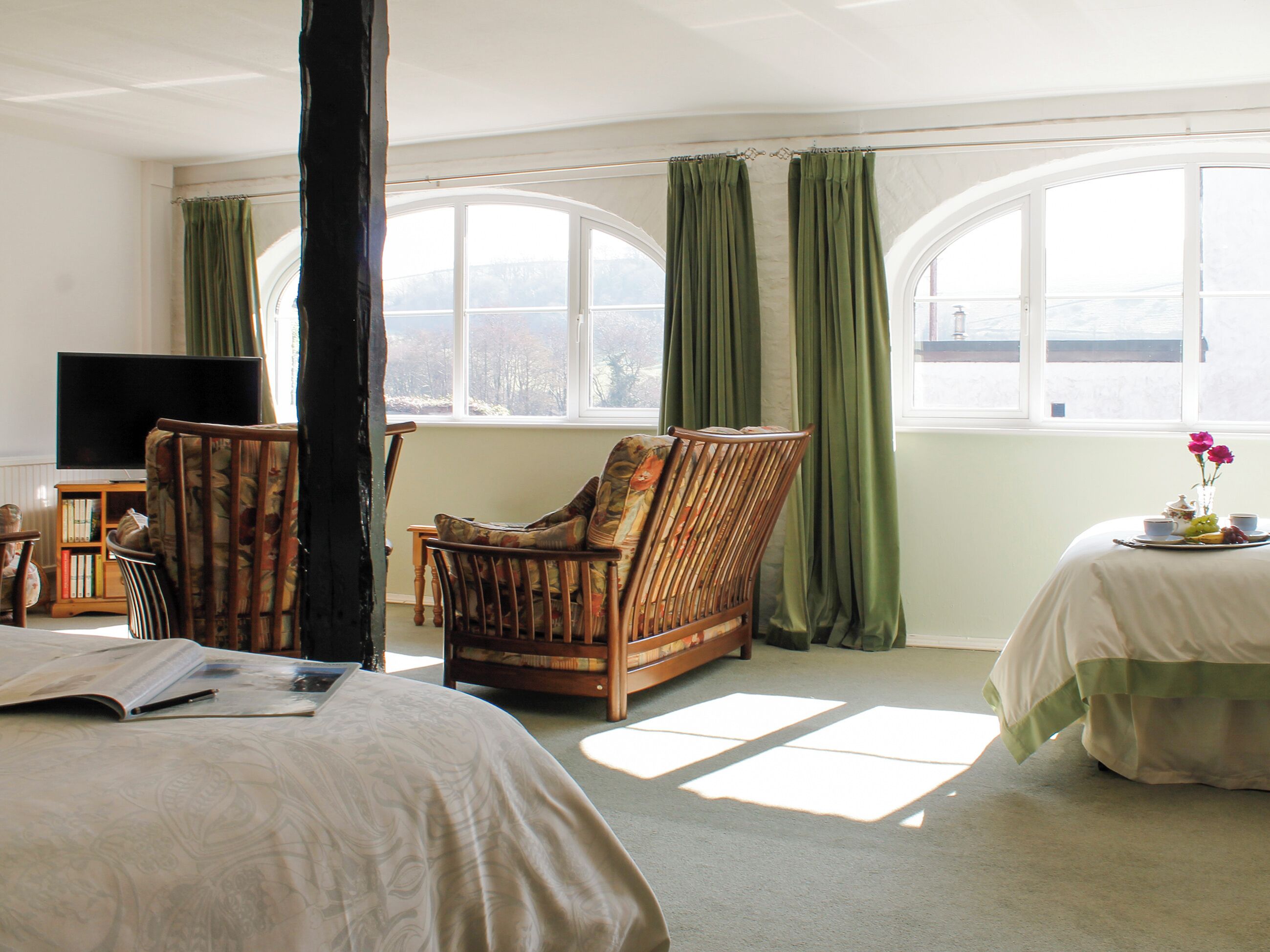A room with a bed, a chair, a television, a window with curtains, and a table with a vase of flowers.