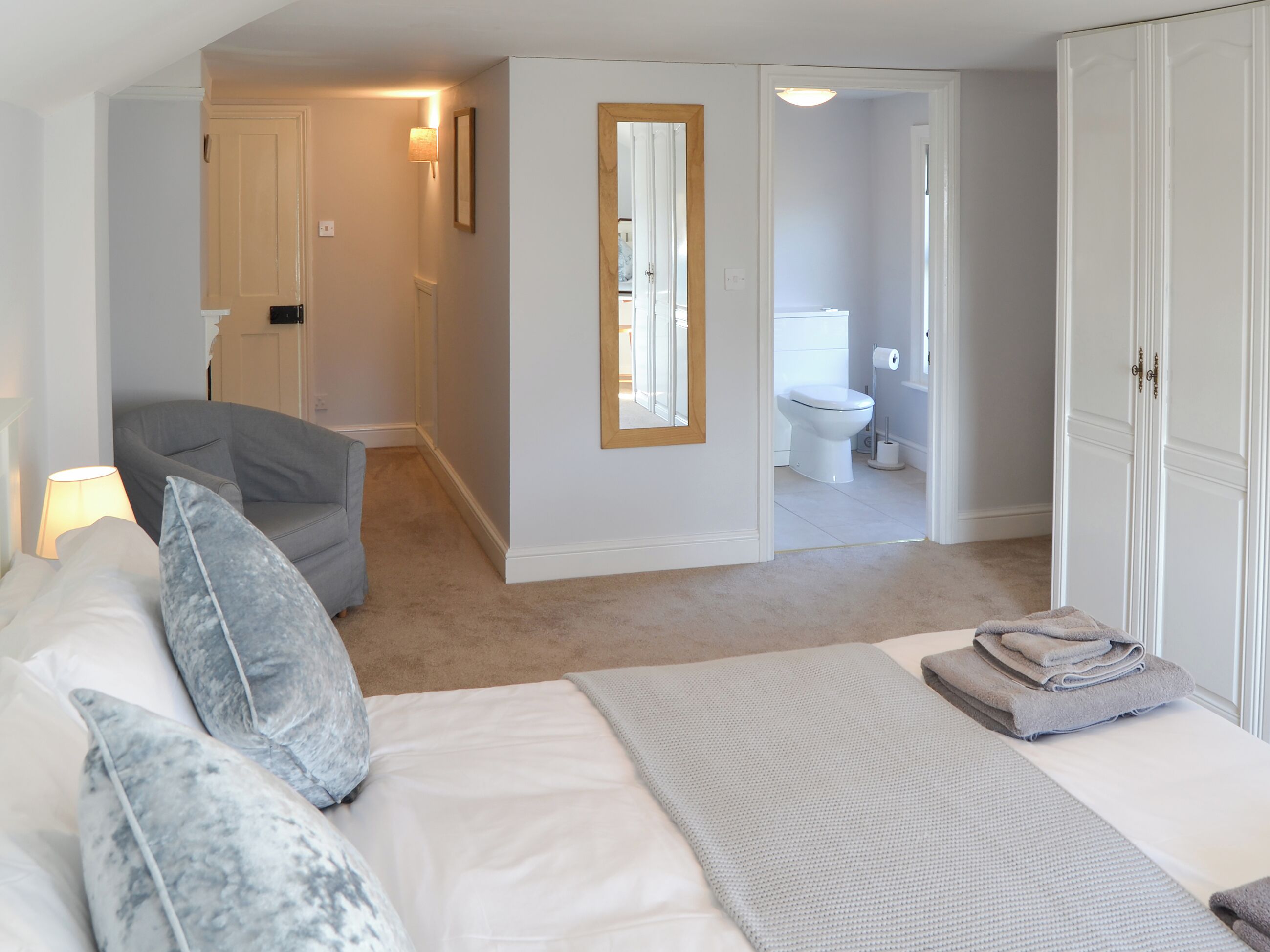 A bedroom with a bed, a chair, a bedside table with a lamp, and a bathroom visible through an open door.
