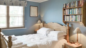 Una camera da letto con un letto in legno, biancheria bianca, un comodino con una lampada, una libreria e una finestra con persiane.