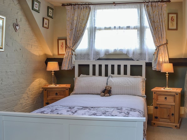 Une chambre à coucher avec un lit blanc, deux tables de chevet, un ours en peluche et une fenêtre ornée de rideaux à motifs floraux.