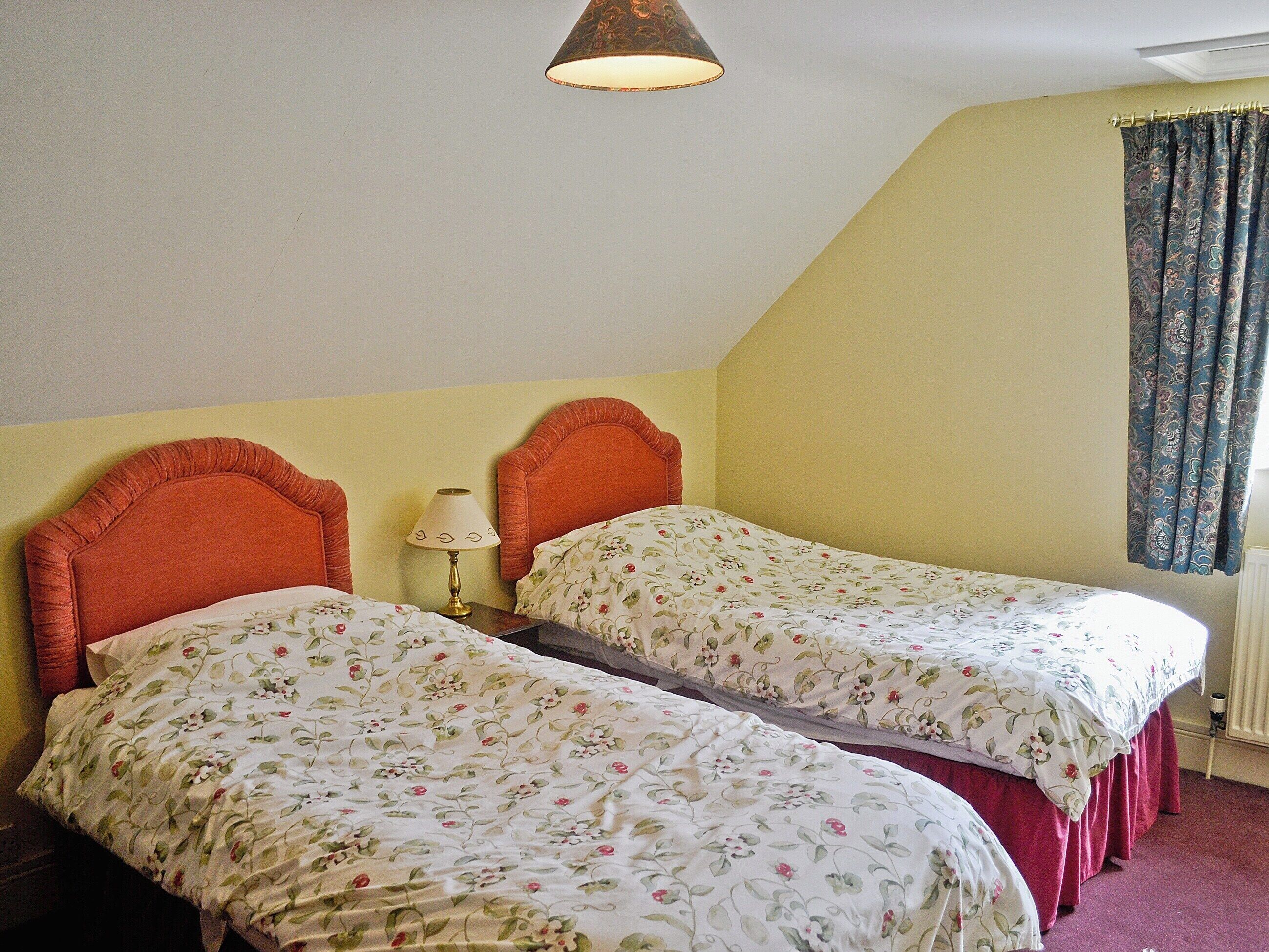 Une chambre avec deux lits simples, chacun agrémenté d’une literie à motifs floraux, une table de chevet avec une lampe et une fenêtre avec des rideaux.
