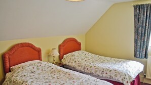 A room with two single beds, each with floral bedding, a bedside table with a lamp, and a window with curtains.