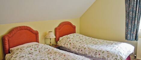 A room with two single beds, each with floral bedding, a bedside table with a lamp, and a window with curtains.