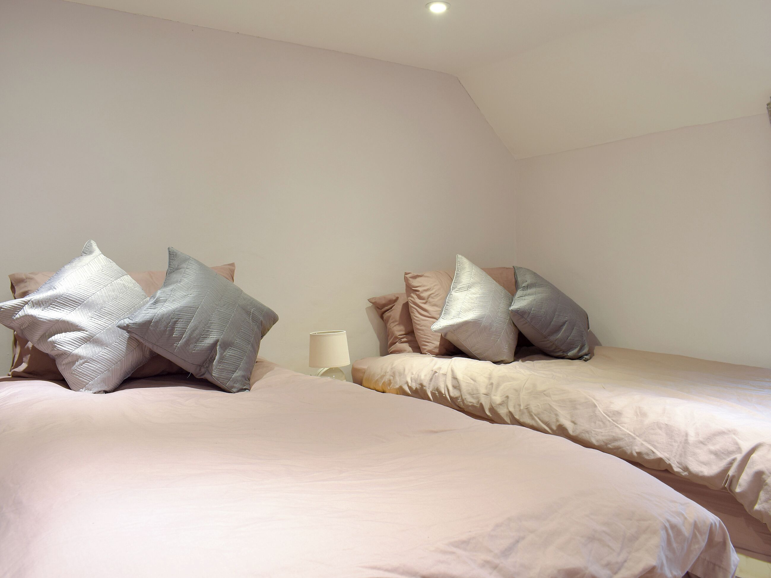 Zwei Betten mit Kissen und ein Nachttisch mit Lampe in einem Zimmer mit weißen Wänden und schrägem Dach.