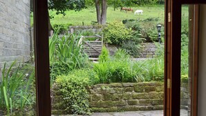 Un chemin de pierres mène à un jardin luxuriant, agrémenté d’une porte en bois, d’un mur de pierres et offrant une vue sur des chevaux qui paissent au loin.