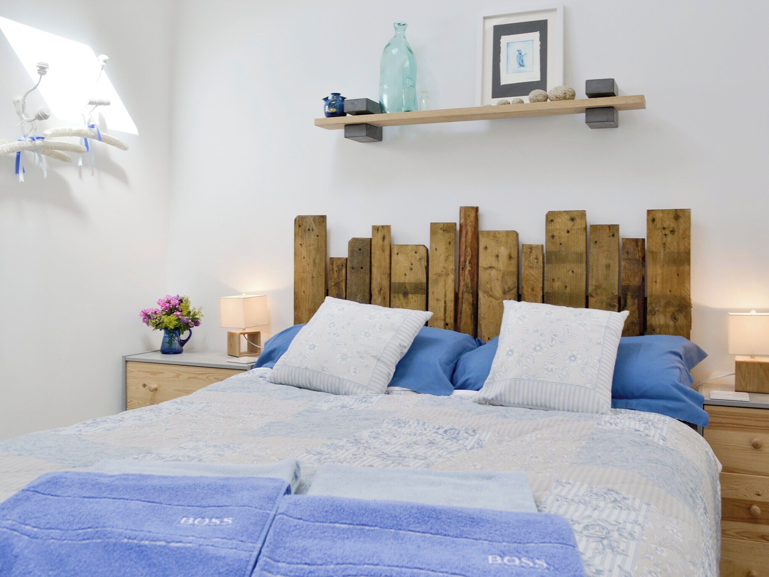 A bedroom with a wooden headboard, a bed with blue and white bedding, a nightstand with a lamp, and a shelf with decorative items.