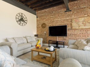 A living room with a brick wall, a clock, a flat-screen TV, a coffee table with fruits, and two sofas.