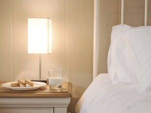 A neatly made bed with white linens, a bedside table with a plate of cookies and a glass of milk, and a lamp providing soft lighting.