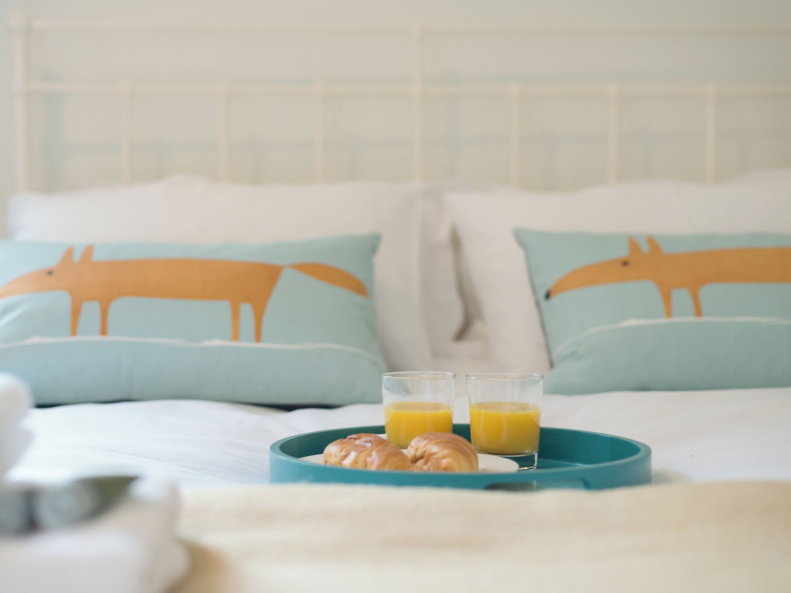 A bed with light blue pillows featuring animal prints, a teal tray with croissants and orange juice, and a white headboard.