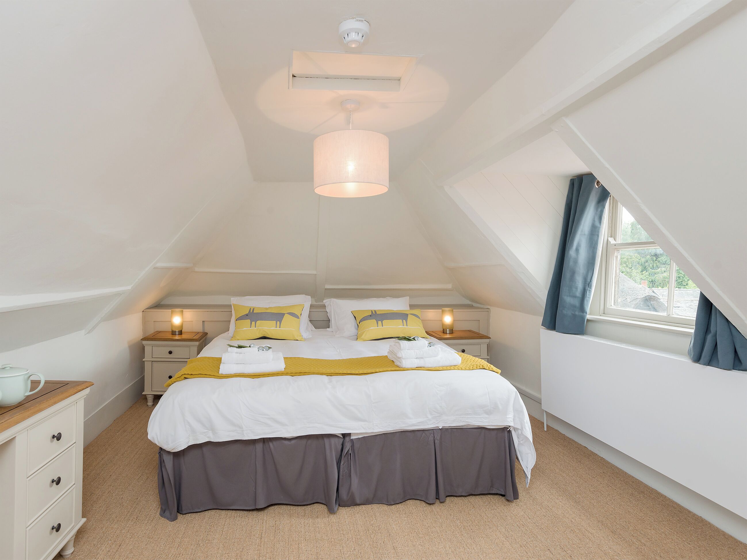 A bedroom with a bed, bedside tables, a hanging light fixture, a window with curtains, and a ceiling light.